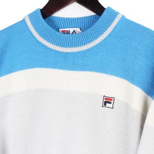 Puede incluir: Un su&eacute;ter vintage de cuello redondo en azul, blanco y gris. La secci&oacute;n superior es azul claro, seguida de una franja blanca y una secci&oacute;n inferior gris. El su&eacute;ter presenta un peque&ntilde;o logotipo en el pecho.