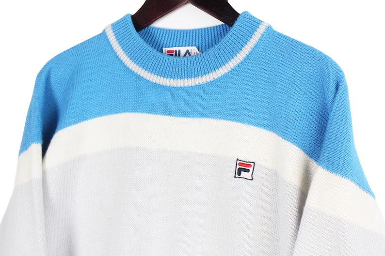 Puede incluir: Un su&eacute;ter vintage de cuello redondo en azul, blanco y gris. La secci&oacute;n superior es azul claro, seguida de una franja blanca y una secci&oacute;n inferior gris. El su&eacute;ter presenta un peque&ntilde;o logotipo en el pecho.