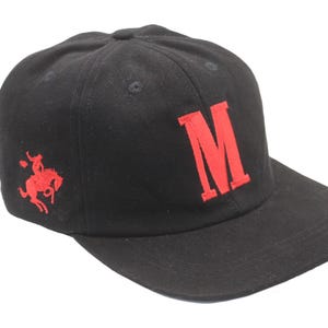 Könnte beinhalten: Schwarze Baseballkappe mit gebogenem Schirm. Die Kappe hat ein großes, rot gesticktes "M" auf der Vorderseite und ein rot gesticktes Bild eines Reiters auf einem Pferd an der Seite. Sie hat einen dunkelbraunen, verstellbaren Riemen.
