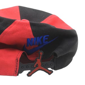 May include: A red and black baseball cap with the Nike logo in blue and the Jordan Jumpman logo. The cap has a snapback closure for adjustable sizing.
