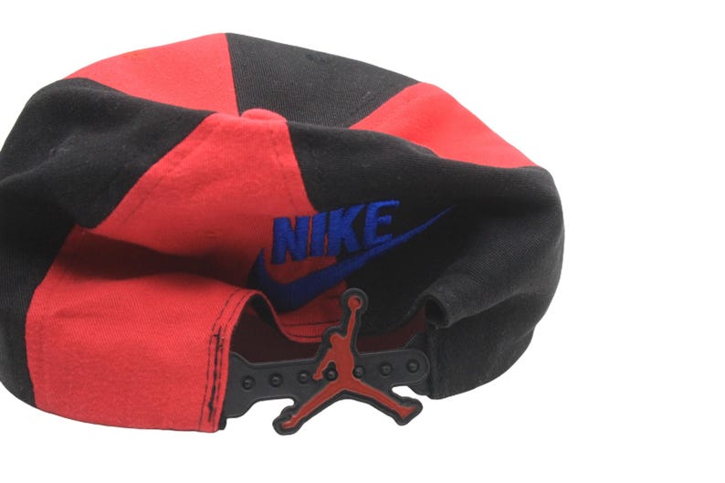 May include: A red and black baseball cap with the Nike logo in blue and the Jordan Jumpman logo. The cap has a snapback closure for adjustable sizing.
