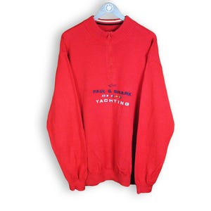 May include: A bright red long-sleeve pullover sweater featuring a quarter-zip mock neck collar. The chest displays the embroidered text PAUL & SHARK YACHTING in blue and white, accompanied by small colorful nautical flag icons above the word YACHTING.