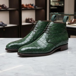 Könnte beinhalten: Ein Paar Stiefeletten in tiefem Smaragdgrün mit glänzender Krokodilprägung. Die Stiefel haben dunkelbraune Schnürsenkel, eine passende dunkelbraune Sohle und eine grüne Zugschlaufe an der Ferse. Das Design umfasst eine abgerundete Zehenpartie.