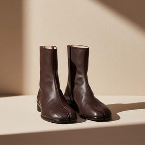 May include: A pair of dark brown leather ankle boots featuring a distinct split-toe design. The boots have a smooth, textured finish, a mid-calf height, and a low, flat black heel. The construction includes a vertical seam running up the front.