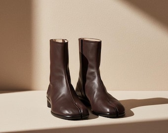 May include: A pair of dark brown leather ankle boots featuring a distinct split-toe design. The boots have a smooth, textured finish, a mid-calf height, and a low, flat black heel. The construction includes a vertical seam running up the front.