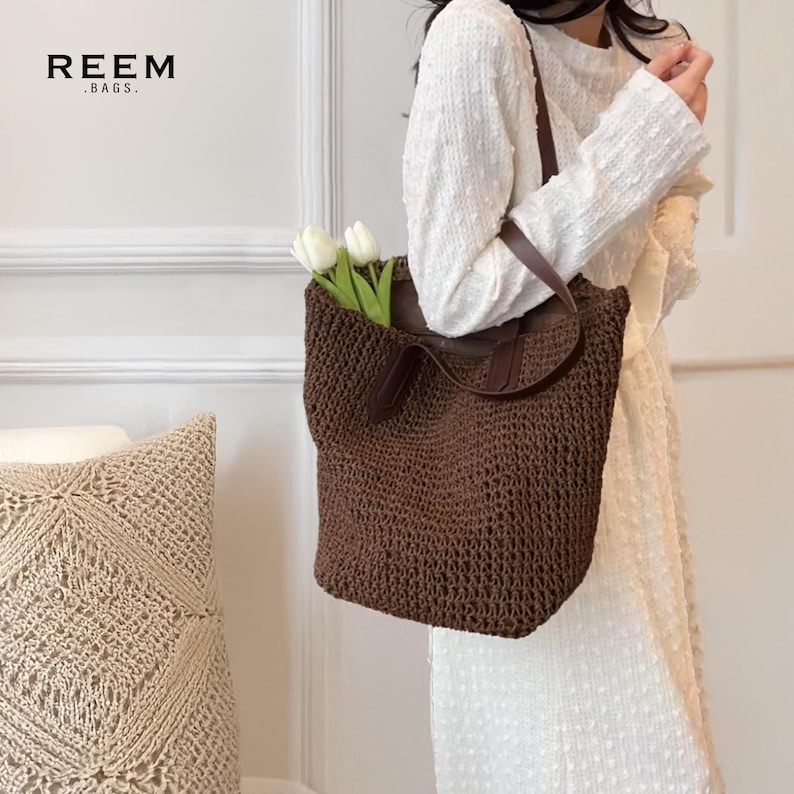May include: A brown woven tote bag with leather handles and a dark interior. The bag is filled with white flowers. The text "REEM .BAGS." is visible in the upper left corner. The bag is a stylish accessory.