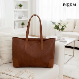 May include: A brown tote bag with a rectangular shape and two handles. The bag has a smooth texture and contrasting brown corners. The brand name "REEM .BAGS." is visible in the top right corner.