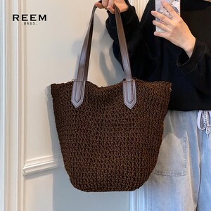 May include: A brown woven tote bag with brown leather handles and light gray accents. The bag has a wide opening and a textured appearance. The brand name "REEM .BAGS." is printed in white at the top left.