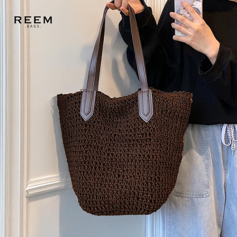 May include: A brown woven tote bag with brown leather handles and light gray accents. The bag has a wide opening and a textured appearance. The brand name "REEM .BAGS." is printed in white at the top left.