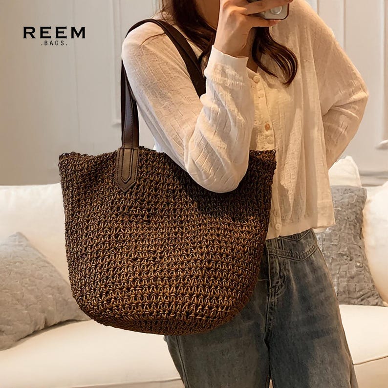 May include: A brown woven tote bag with dark brown leather handles. The bag has a rectangular shape and is being held by a person. The brand name "REEM .BAGS." is printed in the upper left corner of the image.