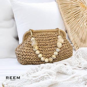 May include: A woven raffia handbag with a beaded handle. The bag is a light tan color with a gold-colored clasp. The handle is made of cream-colored beads. The bag is sitting on a white surface. The brand name "REEM.BAGS" is printed in black.