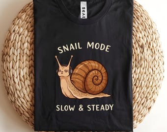 May include: Black t-shirt with the words "SNAIL MODE" and "SLOW & STEADY" printed in white. The shirt features a cartoon snail wearing headphones. The snail has a brown shell and is tan in color.