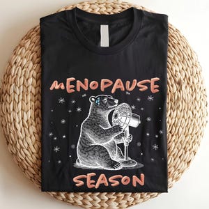 May include: Black t-shirt with the words "MENOPAUSE SEASON" in a coral color. The shirt features a detailed illustration of a bear holding a fan, with snowflakes scattered around the bear.