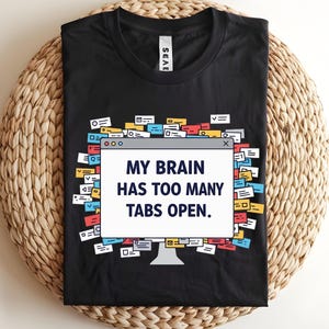 May include: Black t-shirt with the text "MY BRAIN HAS TOO MANY TABS OPEN." The design features a computer monitor graphic surrounded by colorful tabs. The shirt is laid on a woven, circular surface.