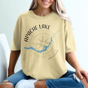 Peut inclure: Un t-shirt à manches courtes jaune pâle avec un motif de boussole noire centré sur la poitrine. Le texte indique APACHE LAKE en arc au-dessus de la boussole et ARIZONA en dessous. Un contour bleu du lac est superposé au motif.