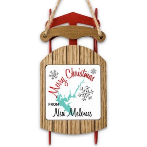 May include: A decorative wooden sled ornament featuring a red metal frame and a wood-grain finish. The center panel displays the text Merry Christmas FROM New Melones in red and black script, accompanied by a teal map shape and silver snowflakes.