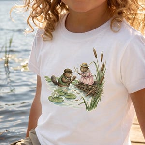 Puede incluir: Camiseta blanca con una ilustración caprichosa de dos castores en un estanque. Uno lleva chaleco y gorra verdes, el otro un vestido y sombrero rosas. La escena incluye nenúfares, juncos y una presa de castor.