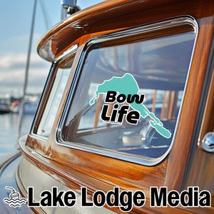 Puede incluir: Un adhesivo con forma de mapa de color cerceta aplicado en la ventana de cristal de una cabina de barco de madera. El adhesivo muestra el texto Bow Life en letras negras negritas con un borde blanco sobre el mapa. Lake Lodge Media aparece abajo.