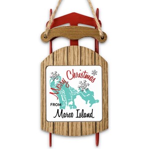 Op de afbeelding: Een decoratieve houten slede-ornament met een rood metalen frame en een houtnerfafwerking. Het midden toont een wit vierkant met een turquoise kaart van Marco Island, twee zilveren sneeuwvlokken en de tekst Merry Christmas FROM Marco Island.