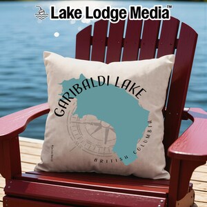May include: A square throw pillow with a cream-colored fabric cover featuring a teal-colored map silhouette of Garibaldi Lake, British Columbia. The design includes black text reading Garibaldi Lake and a faint compass rose graphic behind the map.