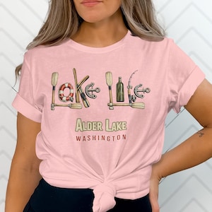 Può includere: T-shirt rosa chiaro con la scritta "Lake Life" formata da illustrazioni nautiche. Il design include un salvagente, un'ancora, una bottiglia e una canna da pesca. Sotto, "Alder Lake Washington".