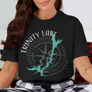 Può includere: Una t-shirt nera a maniche corte con un design grafico che raffigura una rosa dei venti e il contorno del Trinity Lake, in California. Il testo Trinity Lake è stampato in bianco sopra la bussola, con California stampato in bianco sotto.