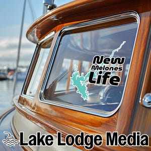 May include: A vinyl decal applied to a boat window featuring a light blue map outline of New Melones Lake and the white text New Melones Life with a black outline. The decal is positioned on the glass of a polished wooden boat cabin frame.