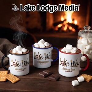 Puede incluir: Tres tazas blancas de estilo esmaltado rellenas de chocolate caliente y nubes. Cada taza muestra el texto Lake Life, Abraham Lake, Alberta, e ilustraciones de remos, un salvavidas y una linterna. Las tazas tienen asas negra, azul y roja.