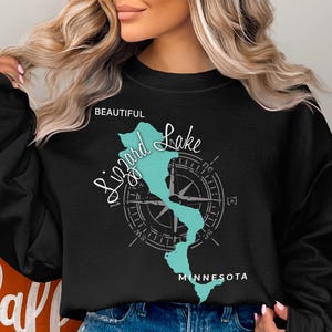 May include: A black long-sleeve sweatshirt featuring a teal map outline of Lizard Lake, Minnesota, overlaid with a white compass graphic. The text "BEAUTIFUL" appears in white above the lake, while "Lizard Lake" is written in white script.