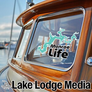 May include: A vinyl decal applied to a boat window featuring a light teal map outline of a lake above the black text Monroe Life. The decal includes the small white text Lake Lodge Media LLC below the main lettering on the glass surface.