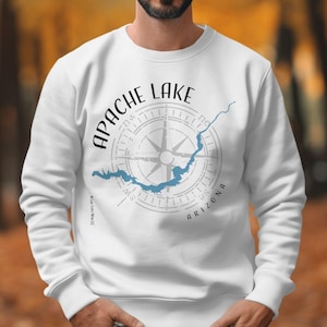 Peut inclure: Un sweat-shirt blanc à col rond avec un motif graphique sur la poitrine. Le design comprend le texte APACHE LAKE en noir, une silhouette bleu clair du lac, un fond en forme de rose des vents grise et le mot ARIZONA en lettres noires.