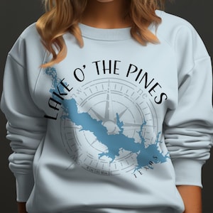 May include: A light blue crewneck sweatshirt featuring a dark blue map silhouette of Lake O' The Pines, Texas. The design includes a compass rose graphic overlaid behind the map and the text "LAKE O' THE PINES" arched across the top in black.