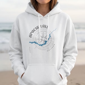 May include: A white hooded sweatshirt featuring a graphic print on the chest. The design includes a grey compass rose, a blue outline of Apache Lake, and the black text "APACHE LAKE" and "ARIZONA" arranged in a circular pattern around the compass.