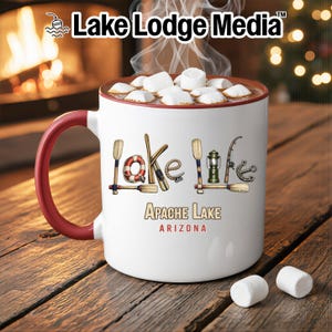 May include: A white ceramic mug with a red handle and rim, filled with hot cocoa and marshmallows. The mug features the text Lake Life, with letters formed by nautical items like oars and a lantern, plus the text Apache Lake Arizona below.