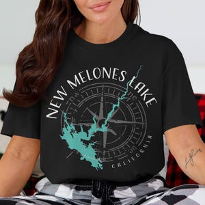 May include: A black short-sleeve t-shirt featuring a graphic print of a compass rose overlaid with a teal map outline of New Melones Lake. The text New Melones Lake is arched above the compass, with the word California printed below the design.