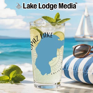 Può includere: Un bicchiere trasparente riempito con una bevanda, cubetti di ghiaccio e fette di lime. Il bicchiere presenta una sagoma blu della mappa del lago Topaz, California, con dettagli a rosa dei venti. Il testo Topaz Lake è stampato sopra.