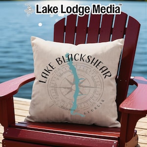 May include: A square off-white throw pillow featuring a black compass rose graphic, a light blue outline of Lake Blackshear, and the black text Lake Blackshear Georgia. The pillow is positioned on a dark red wooden chair with vertical slats.