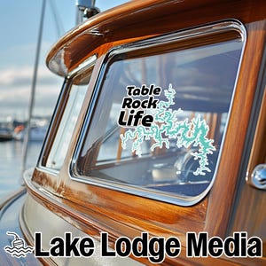 May include: A vinyl decal applied to a boat window featuring the text Table Rock Life in black lettering above a light blue, intricate map outline of a lake. The decal is positioned on the glass of a polished wooden boat cabin structure.