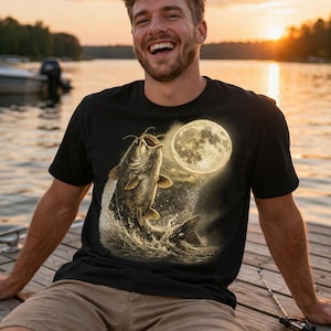 Puede incluir: Camiseta negra con un gráfico de un pez gato saltando hacia una luna llena. La escena está ambientada al atardecer, con una persona sonriendo en un muelle de madera. Se ve una caña de pescar.