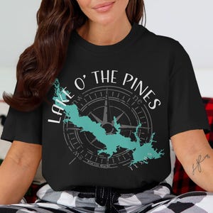 May include: A black short-sleeved t-shirt featuring a graphic design of a teal lake silhouette overlaid on a grey compass rose. The text "LAKE O' THE PINES" is printed in white above the compass, with the word "TEXAS" positioned below the lake.