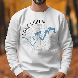 May include: A white crewneck sweatshirt featuring a graphic print of a blue lake outline overlaid on a grey compass rose. The text reads Lake Rabun in black arched lettering above the compass, with Georgia printed in black below the compass.