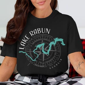 May include: A black short-sleeve t-shirt featuring a graphic design with the text Lake Rabun and Georgia. The design includes a teal outline of the lake superimposed over a grey compass rose with cardinal directions and thin circular lines.
