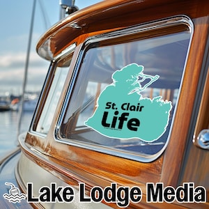 May include: A teal-colored vinyl decal applied to a boat window, featuring a map outline of Lake St. Clair and the black text St. Clair Life. The decal is positioned on the polished wooden frame of the boat cabin window, showing a clear view.