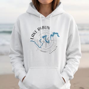 May include: A white hooded sweatshirt featuring a graphic print on the chest. The design includes a grey compass rose overlaid with a blue outline of Lake Rabun, Georgia. The text Lake Rabun is arched above, and Georgia is printed below.