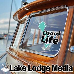 May include: A decal applied to the window of a wooden boat cabin. The decal features a light blue silhouette of a lizard shape alongside the black and white text "Lizard Life". The text "Lake Lodge Media" is visible at the bottom of the image.