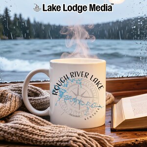 May include: A white ceramic mug featuring a blue map outline of Rough River Lake, Kentucky, overlaid with a compass rose graphic and the text Rough River Lake Kentucky. Steam rises from the mug, which sits near a book and a knit fabric.