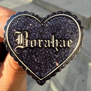 May include: A heart-shaped pin with a glittery dark blue surface and a scalloped edge. The pin features the name "Morahe" in an ornate font. The pin is held up by a hand.