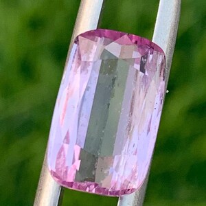 May include: A faceted, rectangular gemstone with a pink and lavender color gradient. The gem is held by silver metal prongs against a blurred green background. The gem's cut reveals multiple facets.