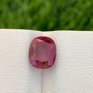 Può includere: Una gemma di rubino sfaccettata, di forma ovale. La gemma è di colore rosso intenso con un aspetto leggermente traslucido. La pietra è posta su una superficie bianca con uno sfondo verde sfocato.