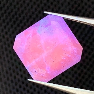 May include: A faceted gemstone with a unique shape, held by tweezers. The gem displays a gradient of pink and purple hues, with internal patterns. The gem's edges are clearly defined, and the overall appearance is polished and luminous.